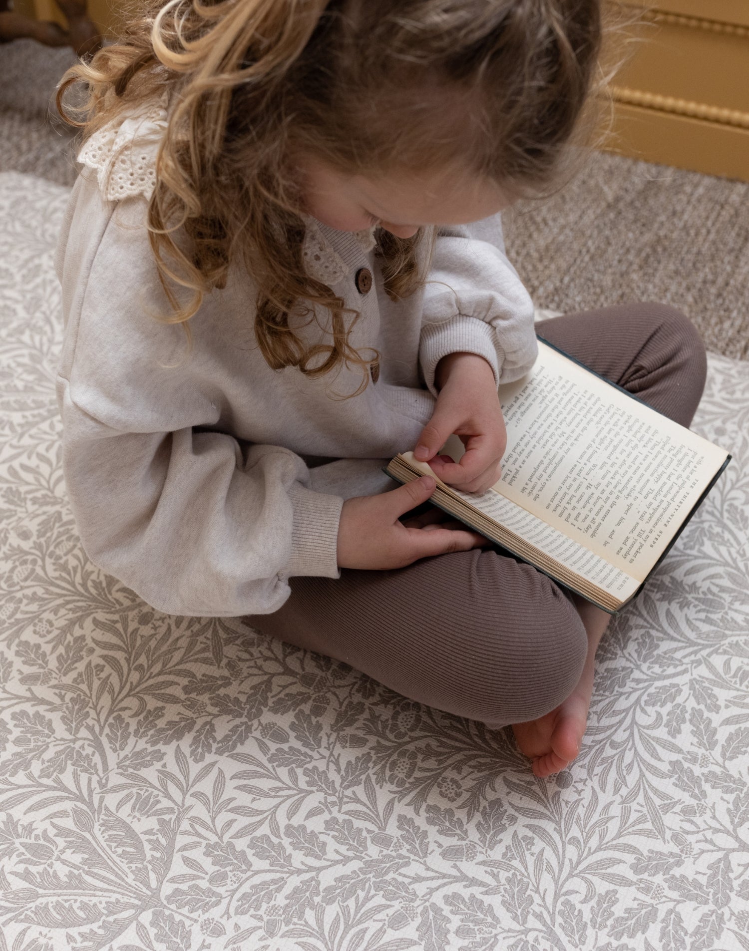 Toddler sits on The Acorn Playmat by Totter + Tumble x Morris & Co. - a luxury padded play mat featuring non-toxic foam and elegant William Morris Acorn motifs.