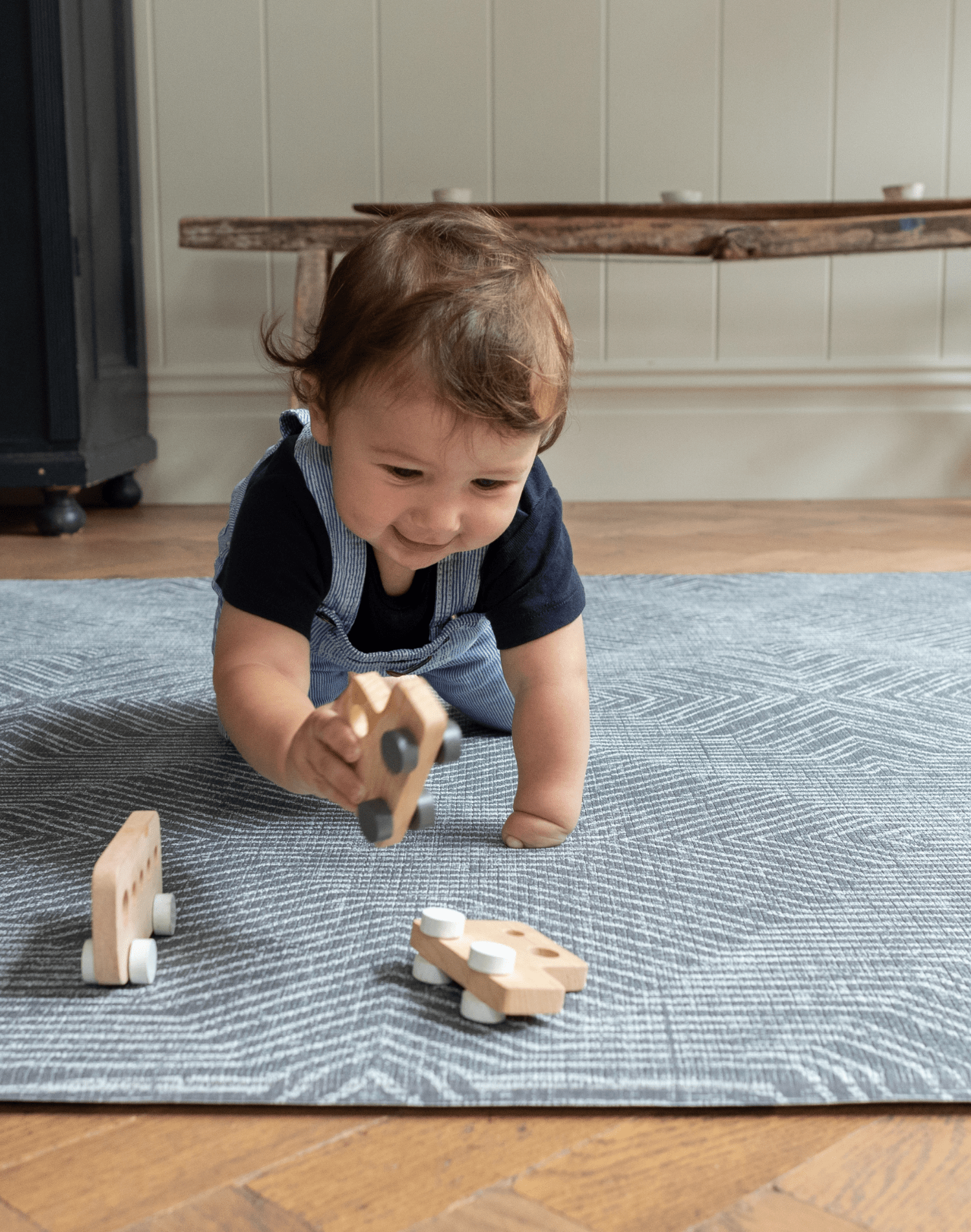 Baby crawls across a textile inspired floor mat playing with a trio of wooden play cars perfect for baby floor play