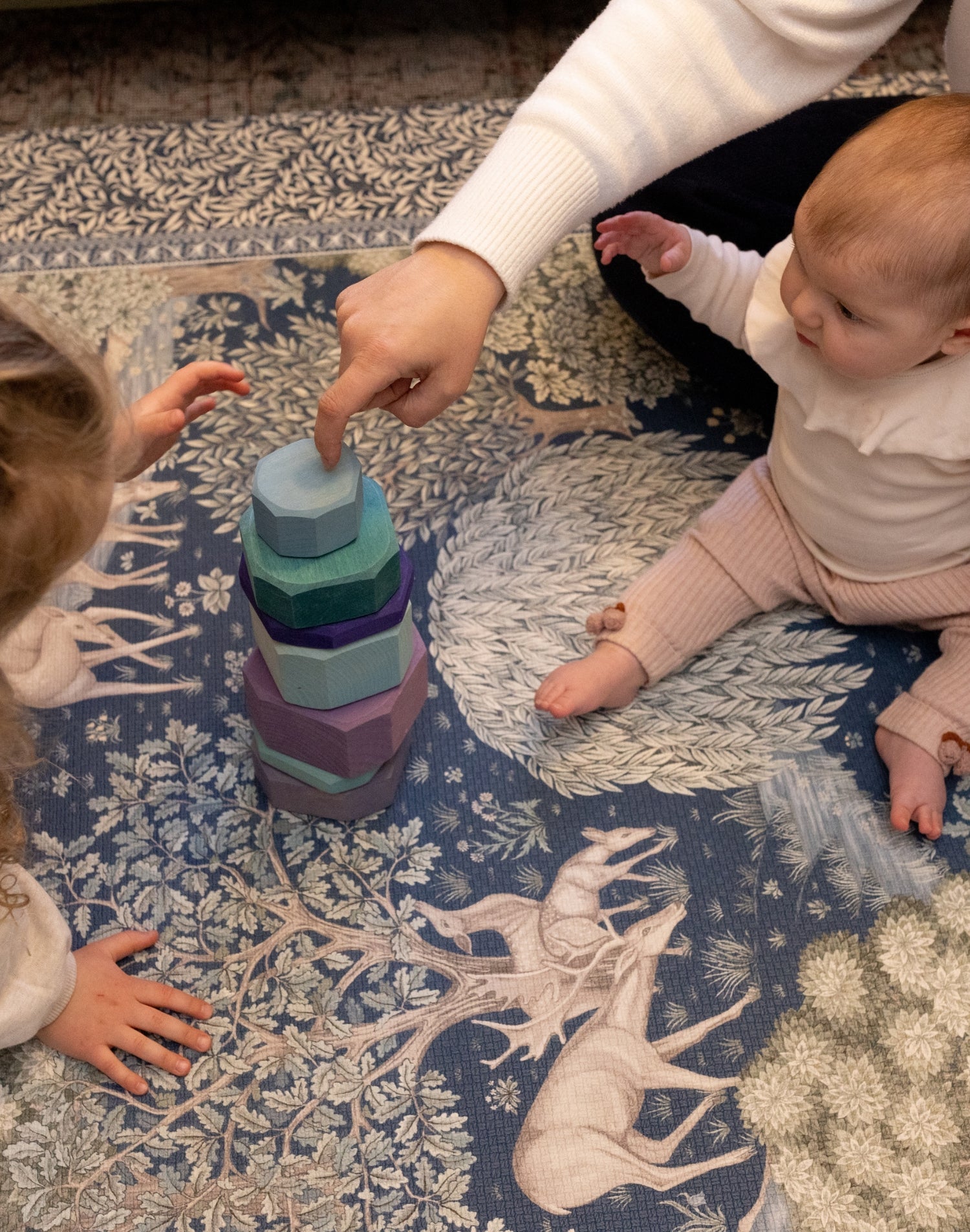 Children play with wooden stacking toys on a botanical-patterned play mat by Totter + Tumble. Safe play on a designer rug.