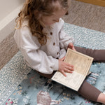 Little girl reads on a William Morris print nursery play mat - practical yet stylish baby product designed for elegant family home aesthetics.