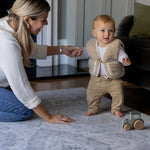 A mother holds the hand of a baby who is learning to walk. The baby is walking on a big soft foam playmat that has a beautiful grey design