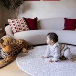 Baby girl sits on the large round playmat the sunda with a delicate leopard print design that complements modern living space the little girl gazes at a large leopard soft toy