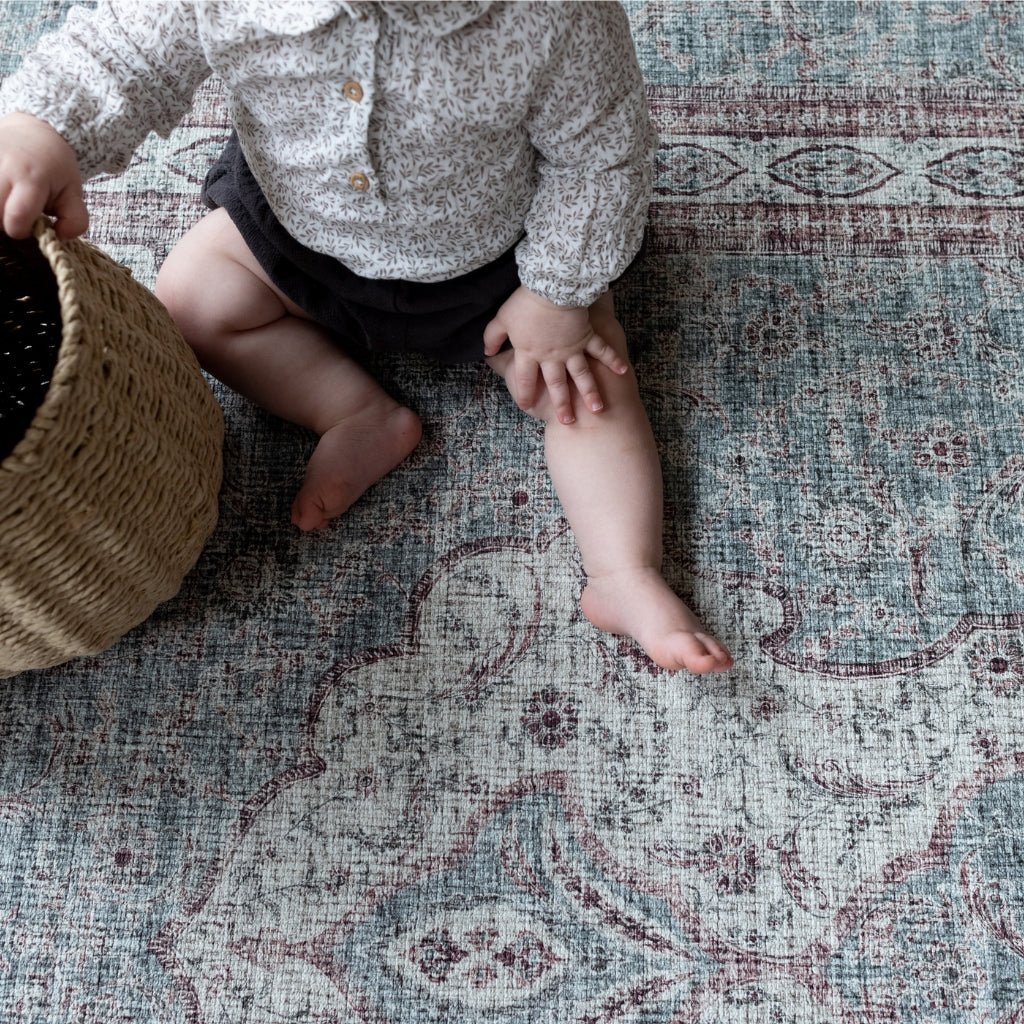 A baby sits on a memory foam playmat rug in deep red and blue-grey tones.