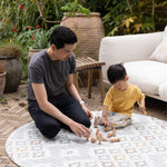 Man and child playing with wooden toys on a round, patterned playmat outdoors.