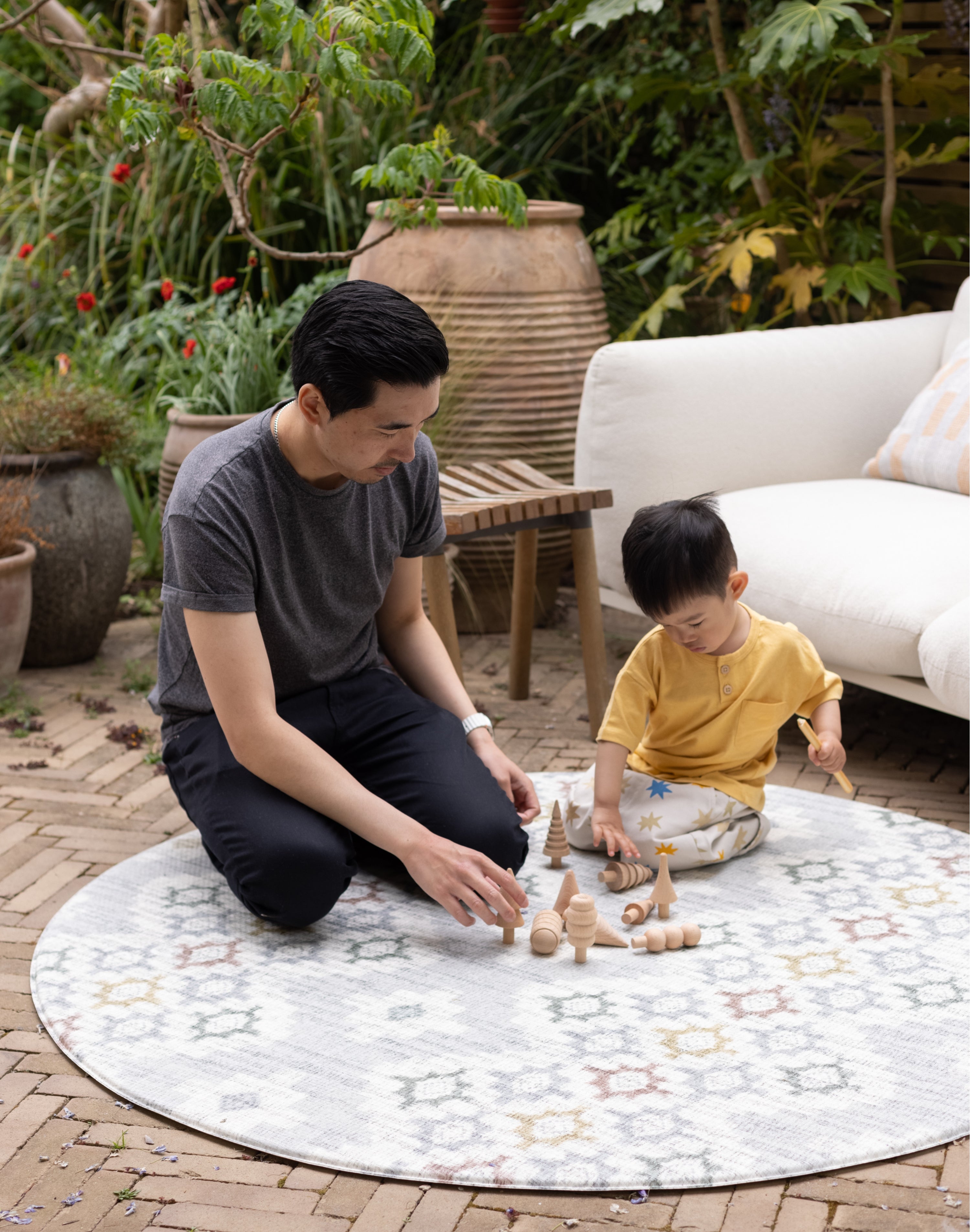 Man and child playing with wooden toys on a round, patterned playmat outdoors.