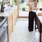 Woman holding a baby in a kitchen with a decorative rug on the floor.