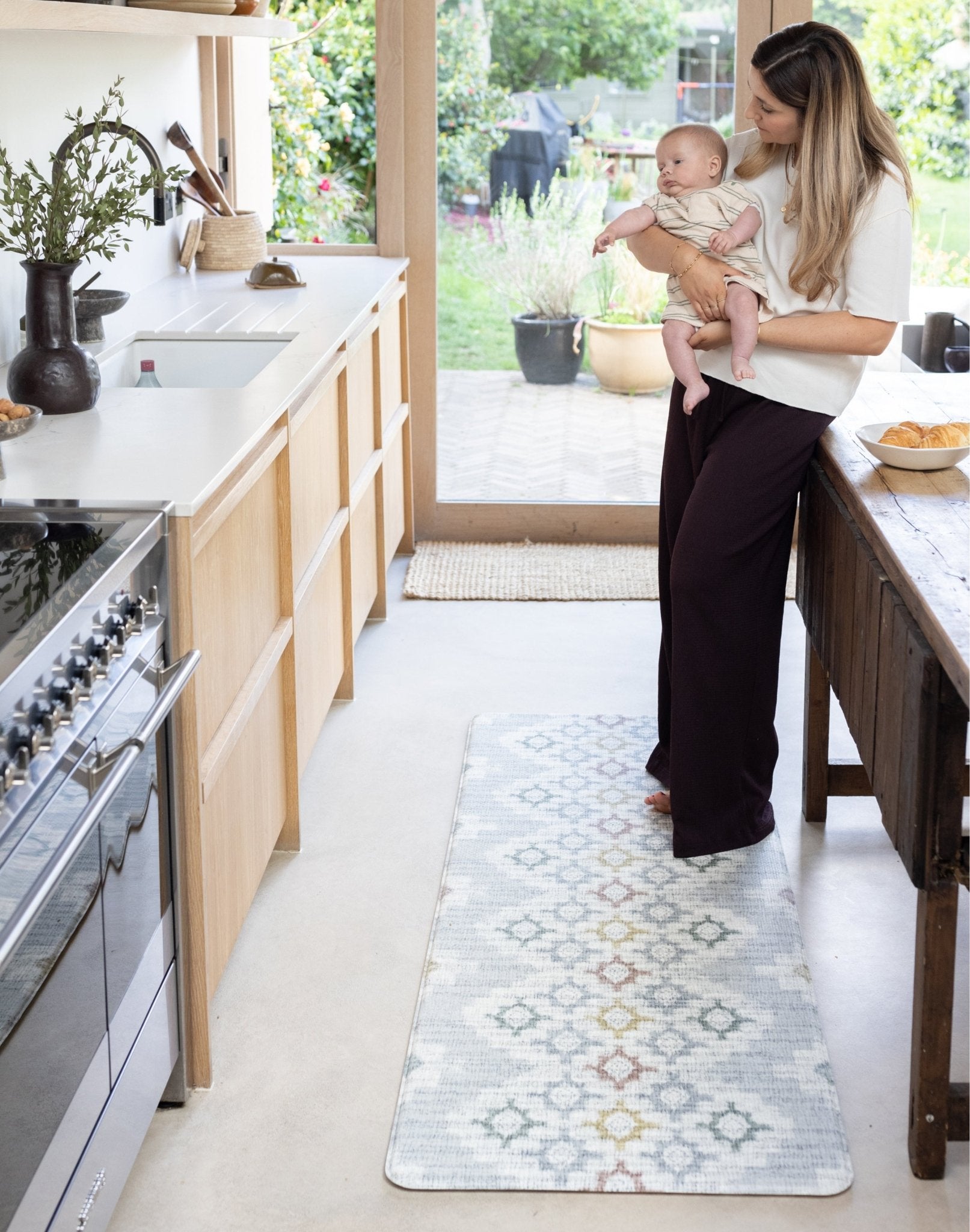 Woman holding a baby in a kitchen with a decorative rug on the floor.