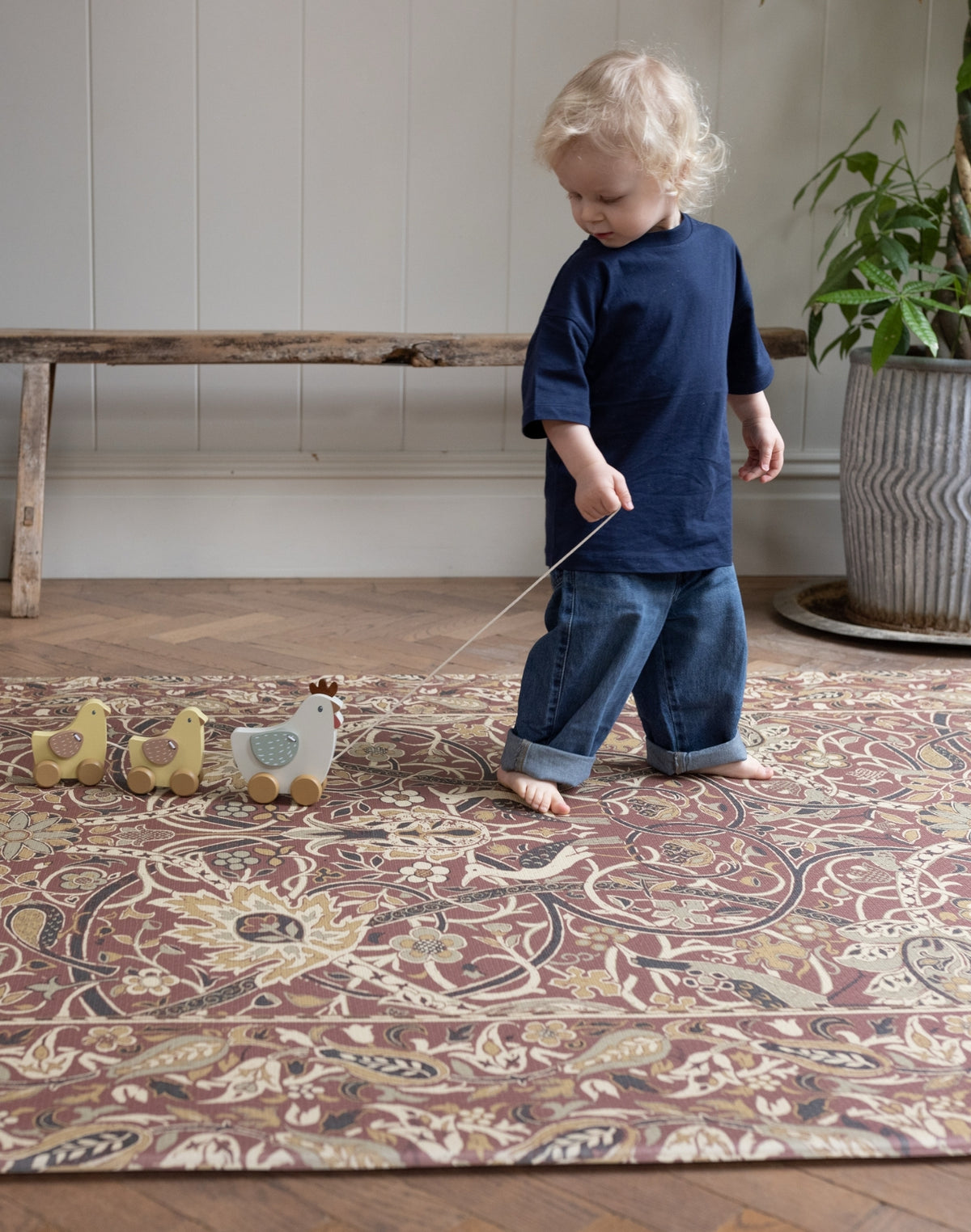 Child playing with toy ducks on a patterned baby play rug for hardwood floors in a room with a wooden bench and plant.