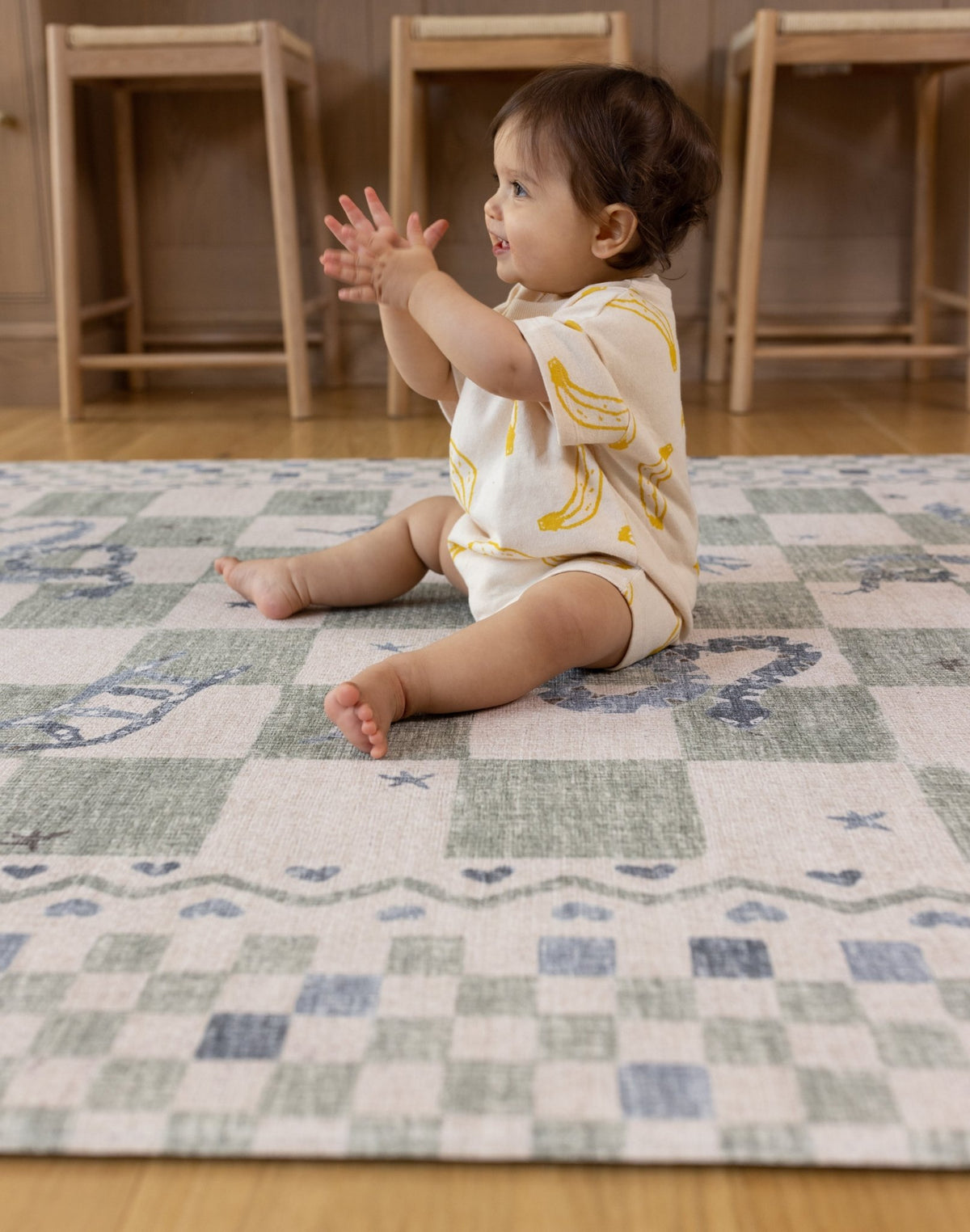 Baby sitting on a modern checkered reversible crawling mat in a room with wooden furniture.