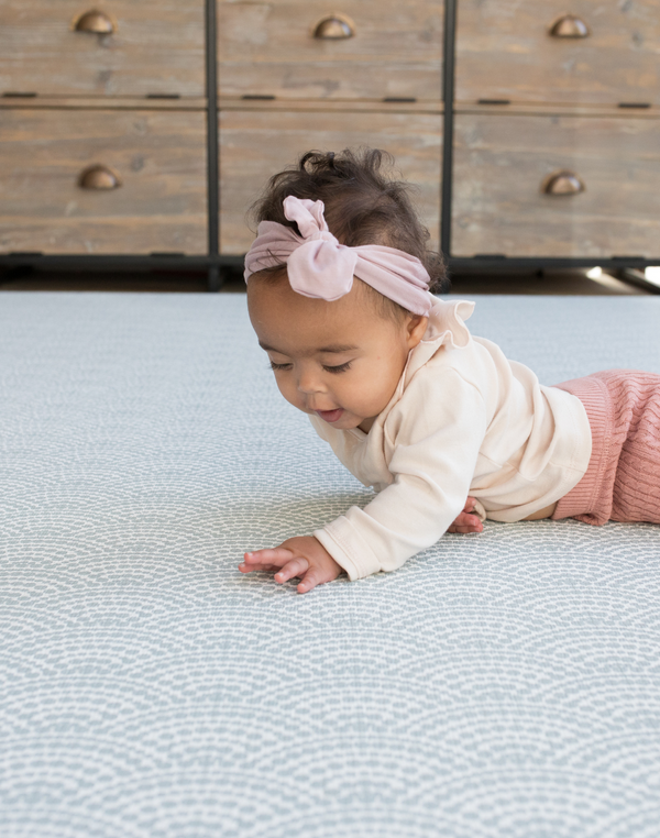Baby with headband lays on protective play mat with modern scalloped design in gentle blue tones 