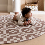 A baby crawls on a round brown patterned playmat. The room has stylish neutral tones
