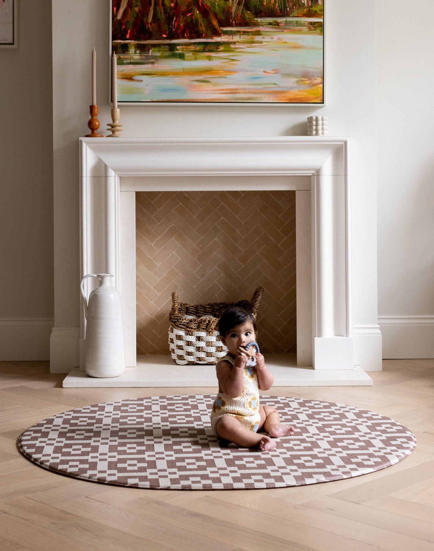 Child sitting on a checkered round baby play mat in a room with a fireplace and painting.