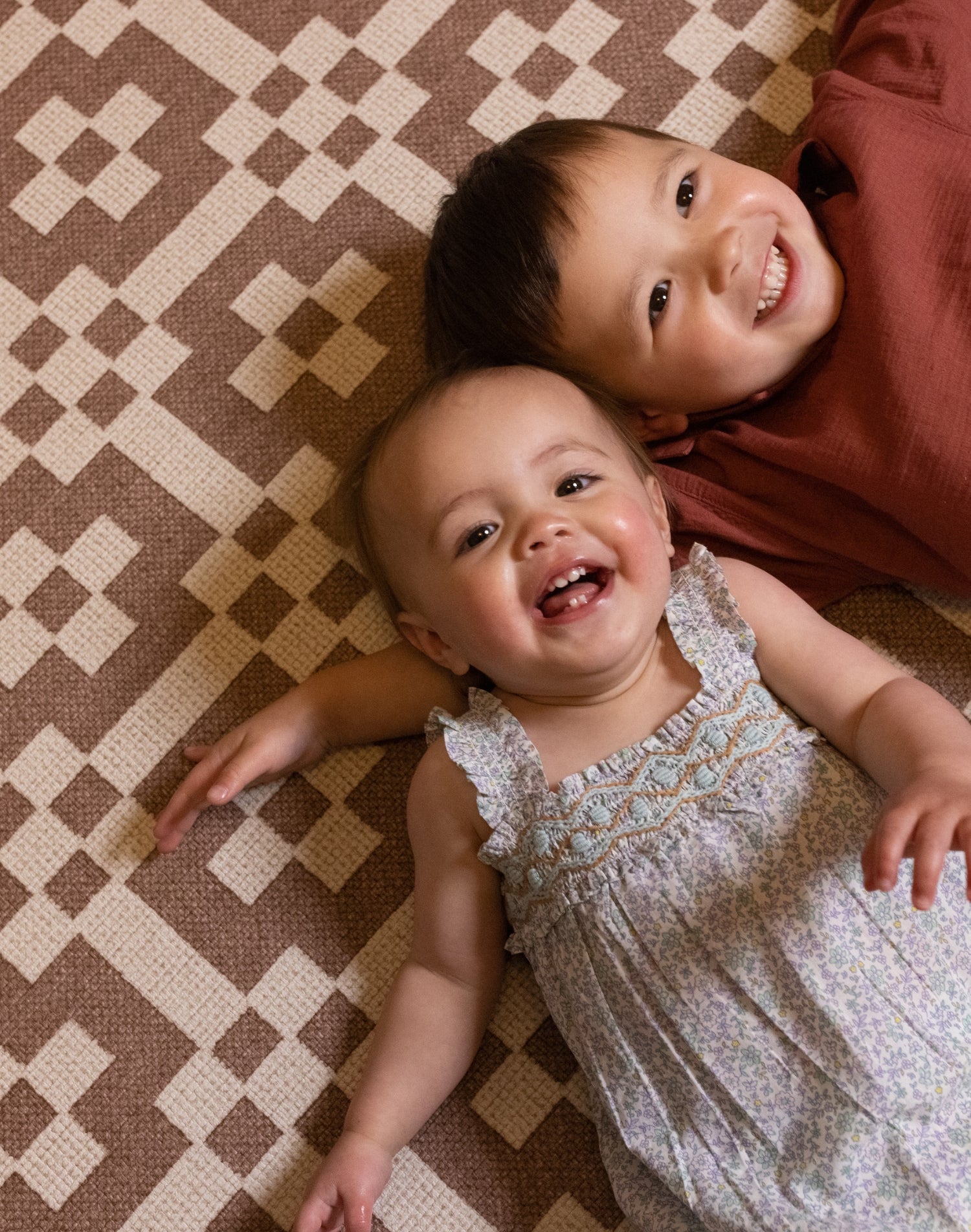 Two children lying down  and smiling on a brown and red patterned playmat from Totter + Tumble