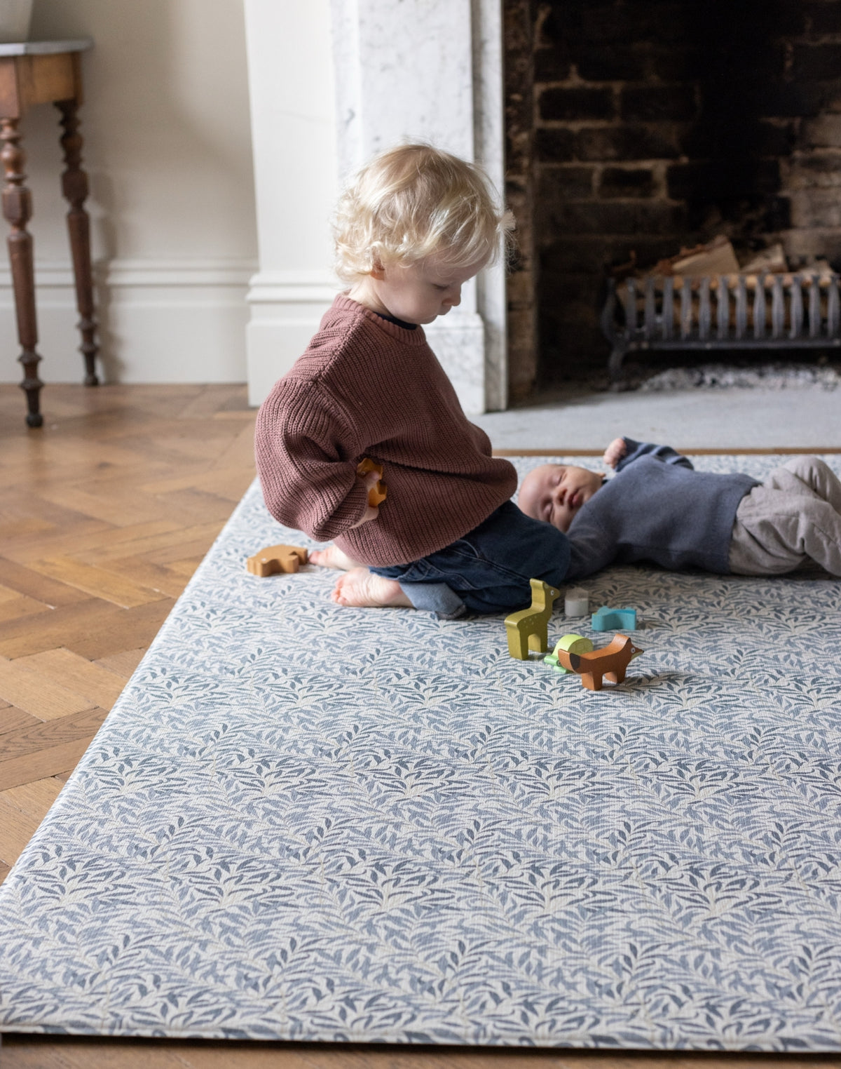Two children playing on a baby safe cushioned and patterned rug in a room with a fireplace.