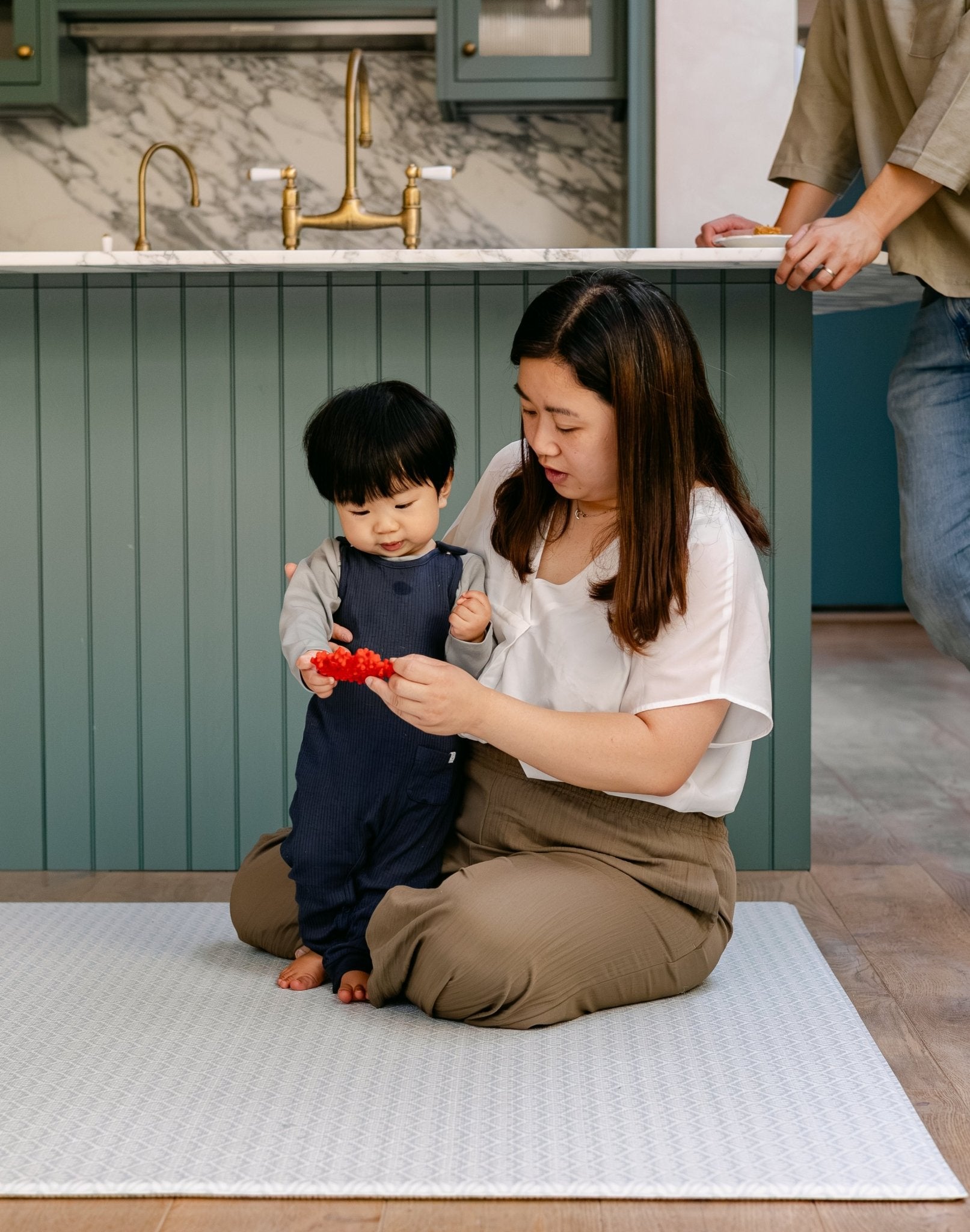 A family sat on the shell design, rug-like beachcomber playmat in a kitchen | SKU: 26 BEACH/GATHER XL, 26 BEACH/GATHER STD, 26 BEACH/GATHER COMP