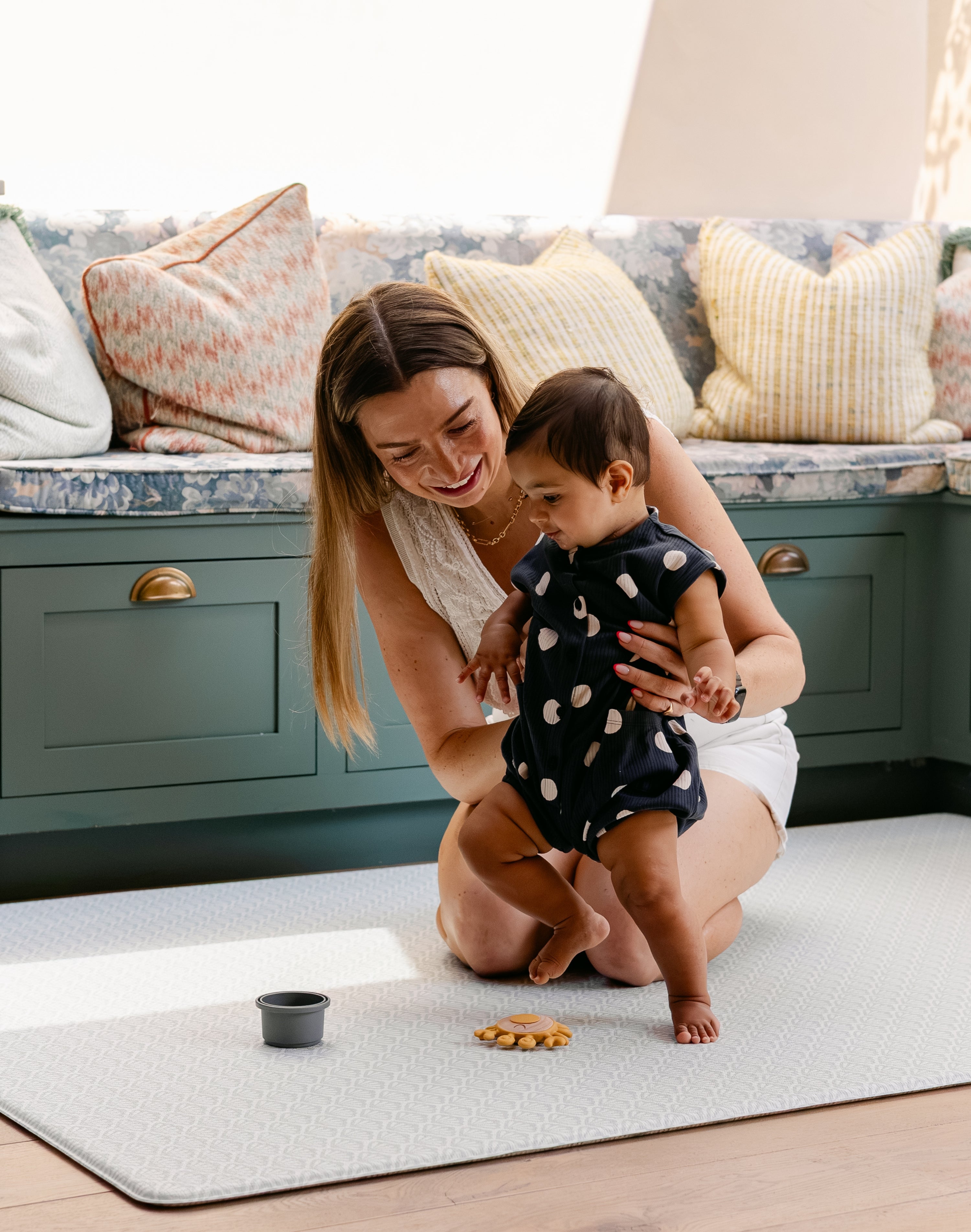 A woman helping her baby take some steps safely and comfortably on a beachcomber totter and tumble playmat. easy to clean, rug-like and non toxic. | SKU: 26 BEACH/GATHER XL, 26 BEACH/GATHER STD, 26 BEACH/GATHER COMP
