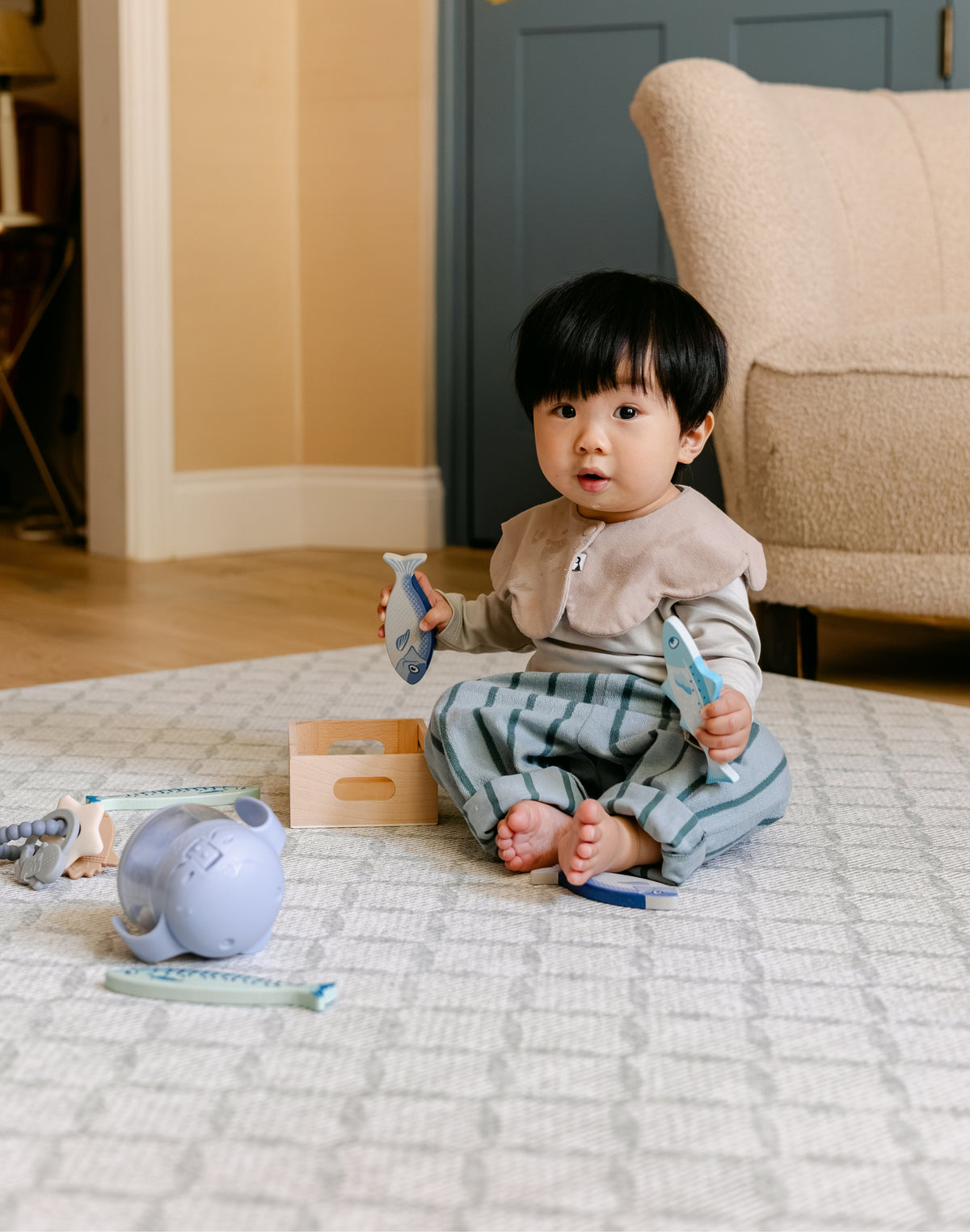 A toddler sits barefoot on a patterned totter and tumble playmat in a living room, holding two light blue toy fish. The child is wearing a beige bib and green striped trousers.