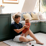 A man sitting on a thick memory foam totter and tumble playmat in a kitchen whilst holding his baby | SKU: 26 BEACH/GATHER XL, 26 BEACH/GATHER STD, 26 BEACH/GATHER COMP