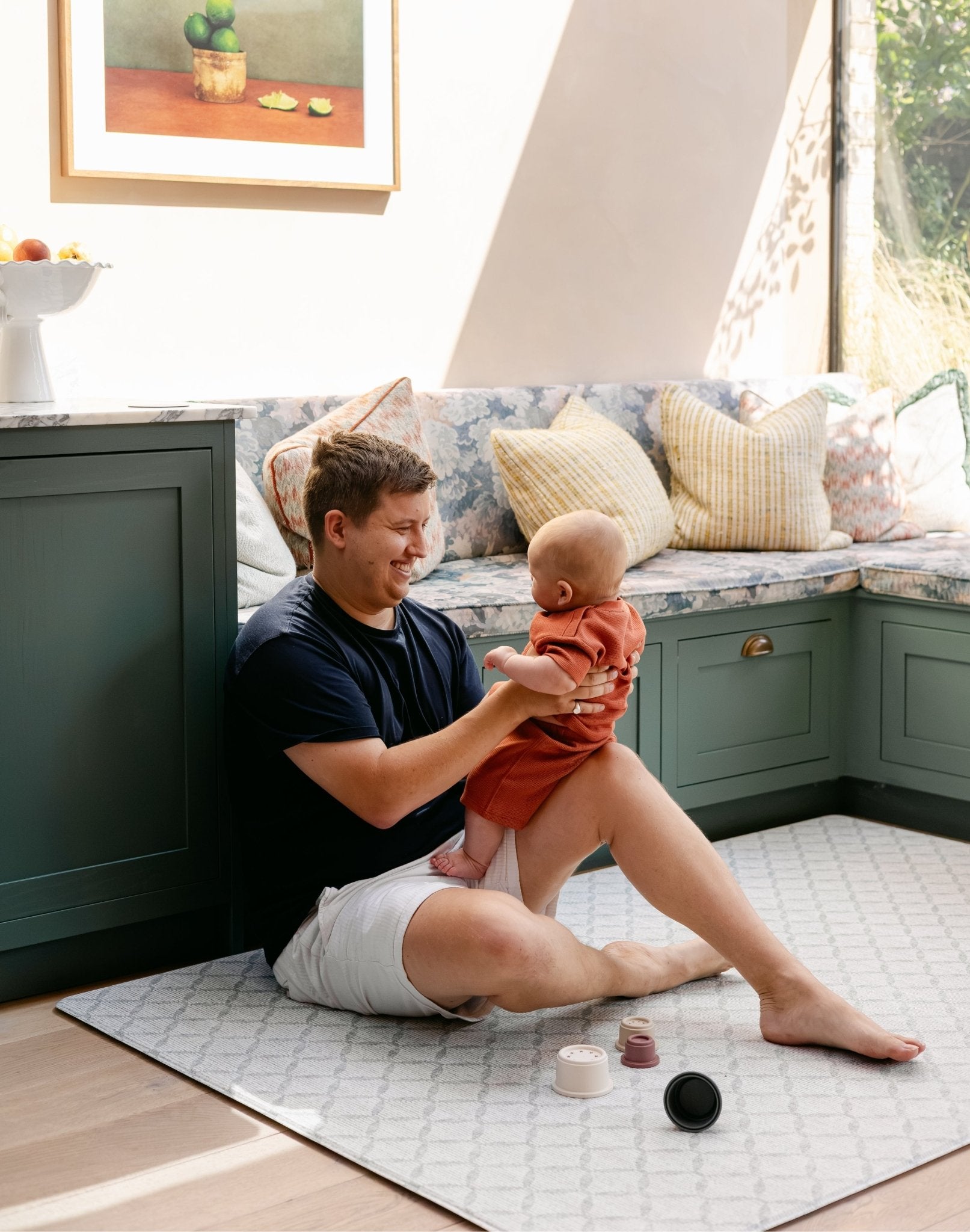 A man sitting on a thick memory foam totter and tumble playmat in a kitchen whilst holding his baby | SKU: 26 BEACH/GATHER XL, 26 BEACH/GATHER STD, 26 BEACH/GATHER COMP