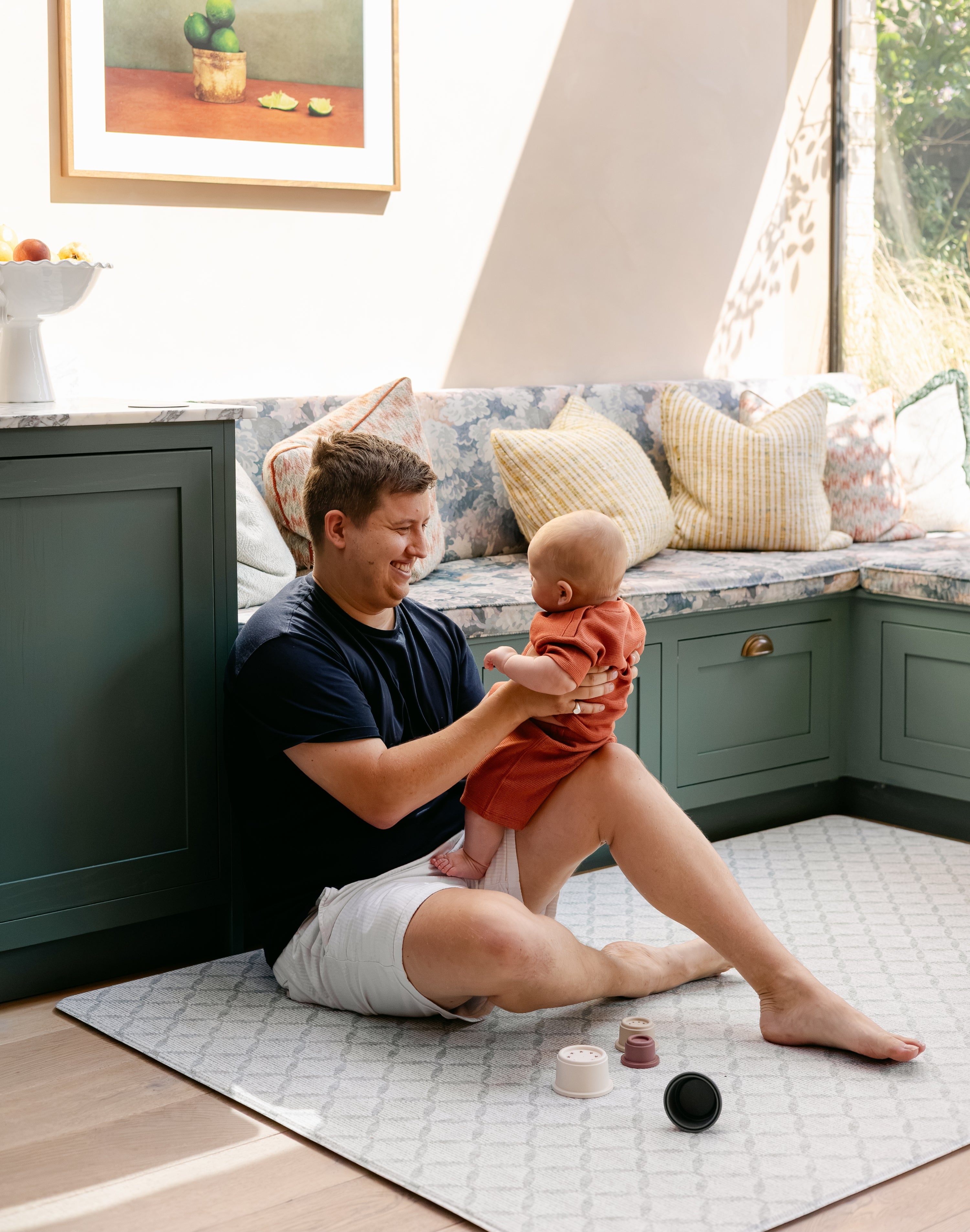 A man sitting on a thick memory foam totter and tumble playmat in a kitchen whilst holding his baby | SKU: 26 BEACH/GATHER XL, 26 BEACH/GATHER STD, 26 BEACH/GATHER COMP